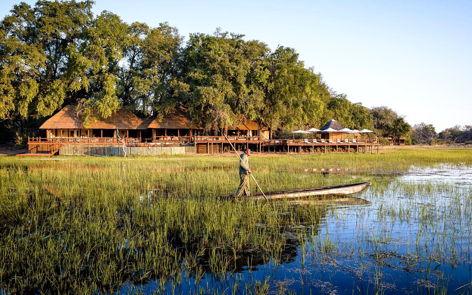 Chief's Camp seen across the water with a guide poling a mokoro past in the foreground in the Moremi