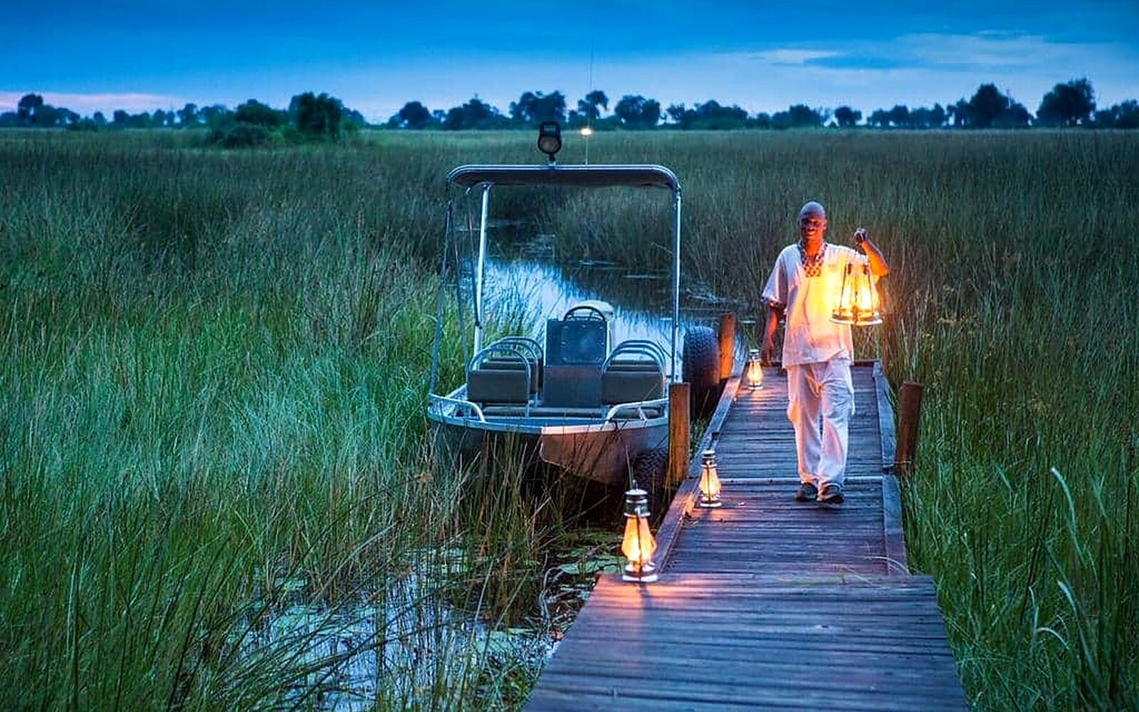 A lantern-lit boardwalk and boat station at Duba Plains at dusk, glowing over the delta reeds