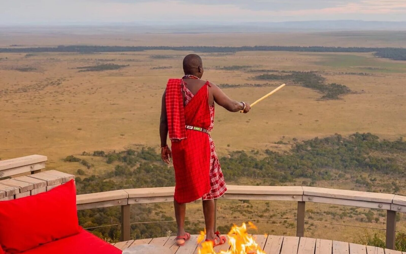 A Maasai guide in a red shukka standing at a fire-pit sundowner deck overlooking the Mara at golden hour