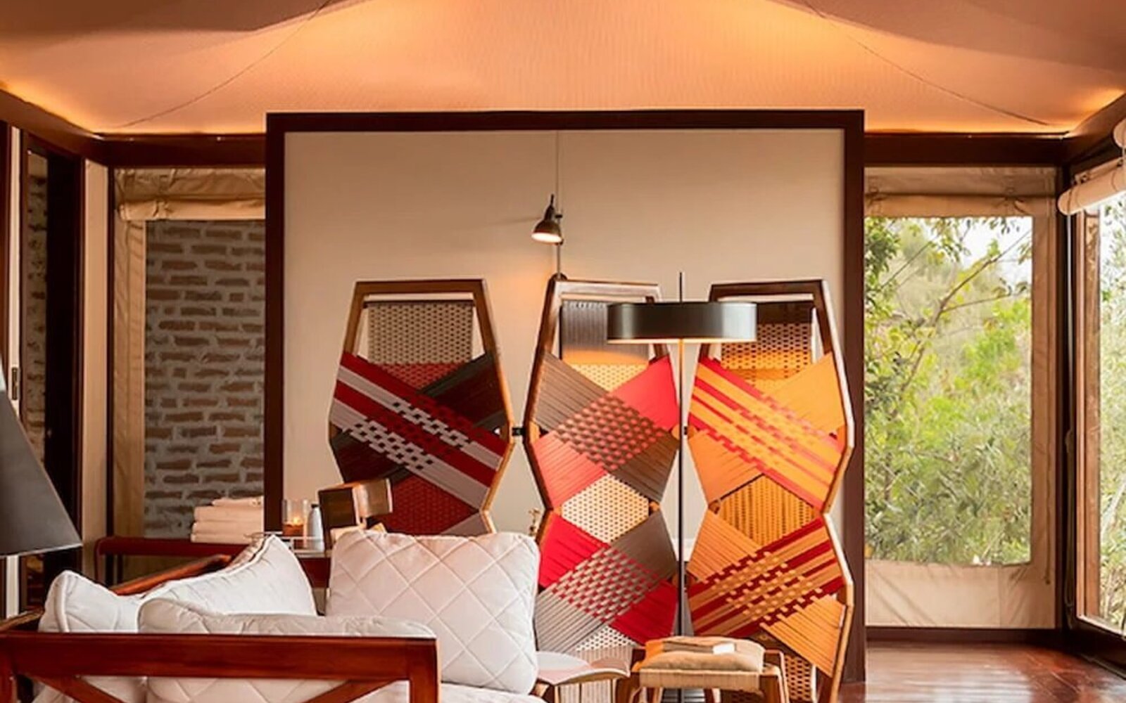 A luxury tented suite bedroom with red Maasai accents and a wall of windows framing the Mara at Angama