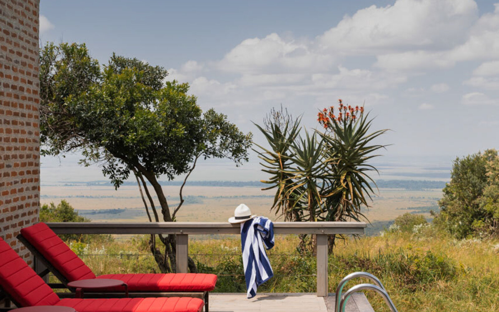 A rim-flow infinity pool and red loungers on a deck looking over the Mara escarpment at Angama