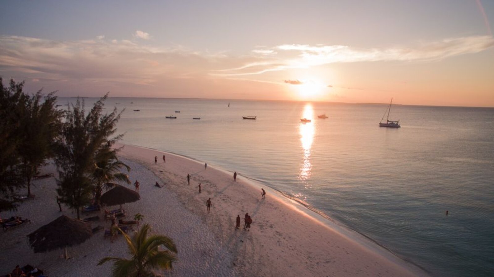 A wide Zanzibar beach at sunset with the sun low over the sea, dhows on the water and a few figures on the sand