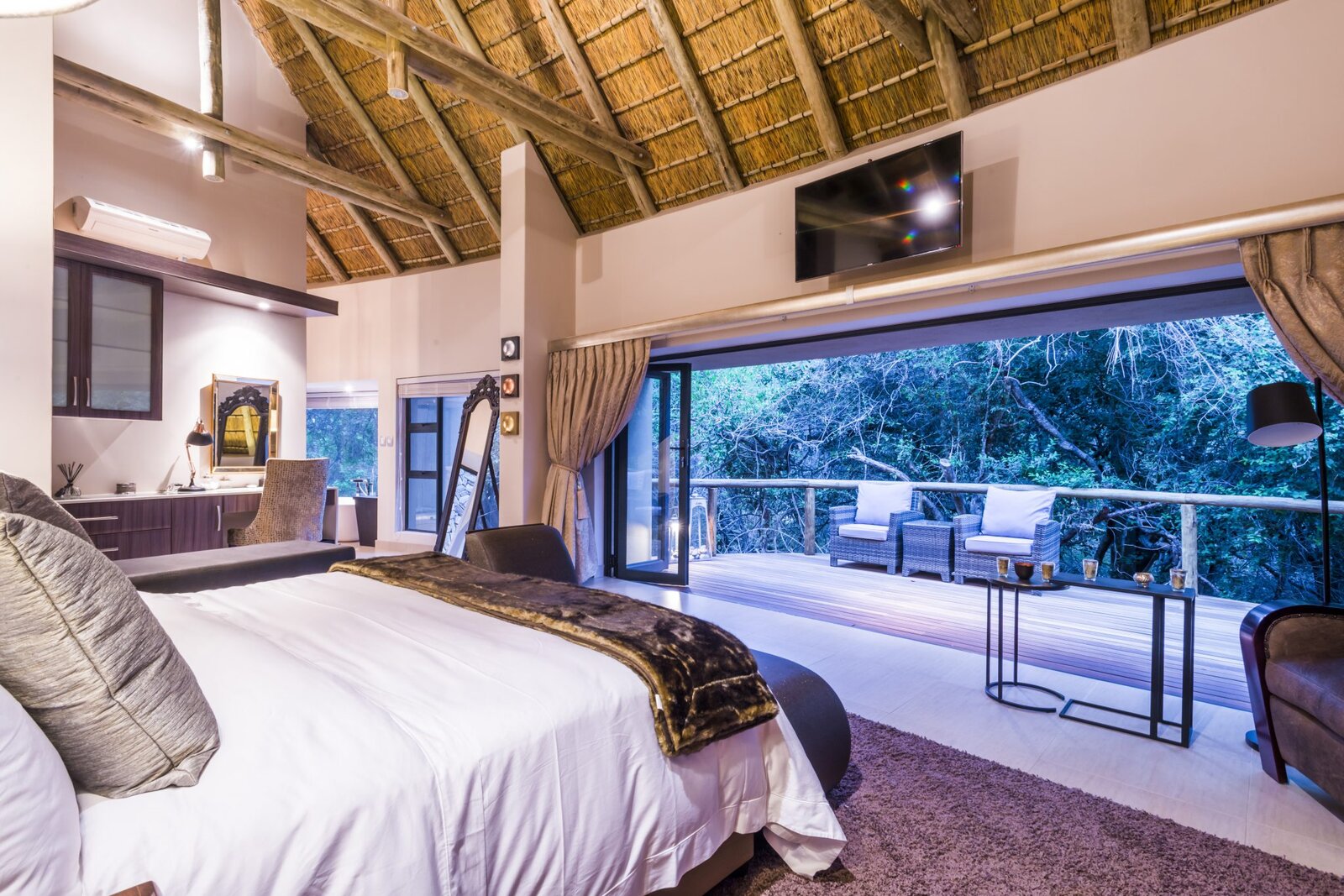 A luxury suite at AM Lodge with a deep bed under a thatched roof opening onto a private deck and the bush at Hoedspruit