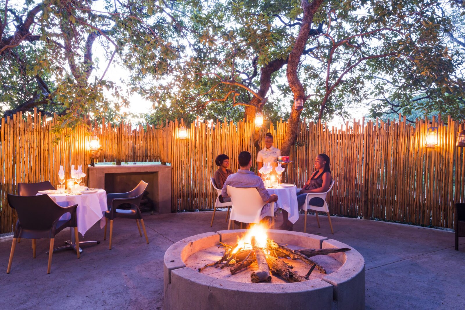Guests at a boma fireside dinner with lanterns and a reed boma at night at AM Lodge, Greater Kruger