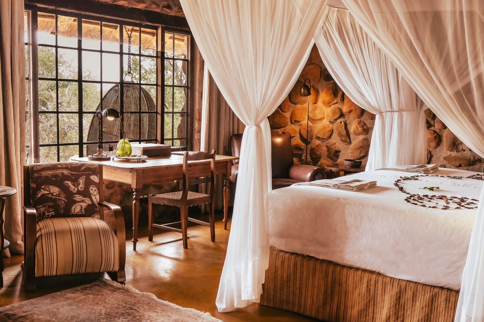 Geiger's Camp suite with four-poster, stone wall and a hanging chair by the window, Timbavati