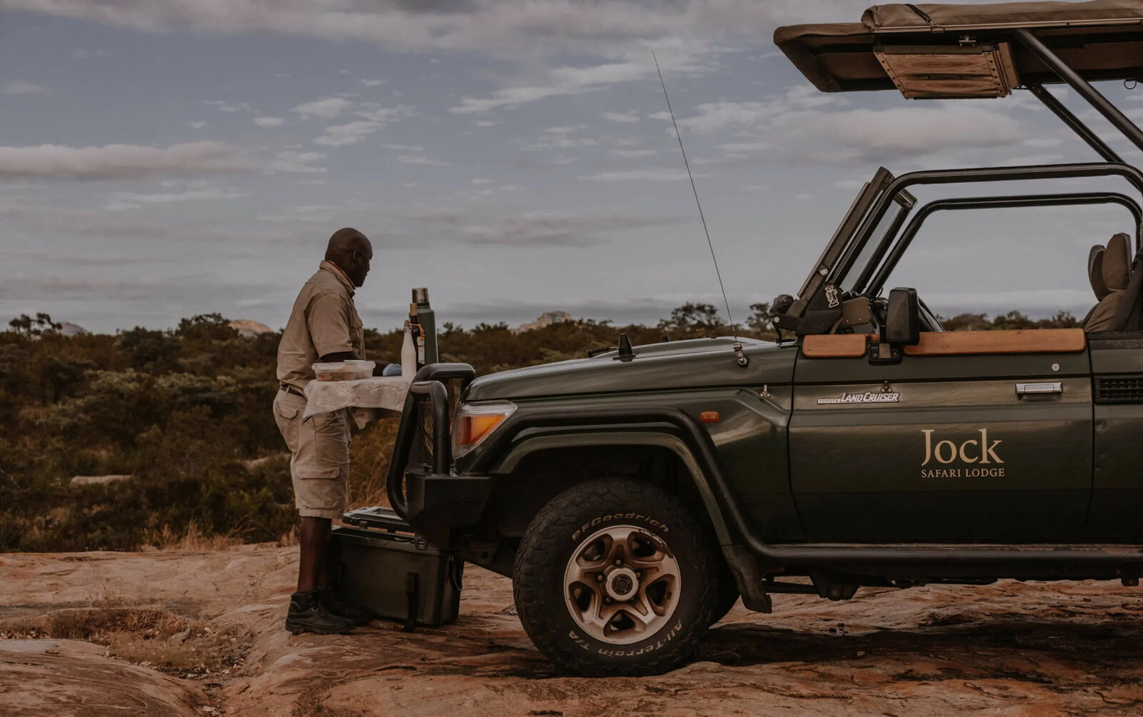A Jock-branded game-drive vehicle at sunset with a guest on the bonnet enjoying sundowners