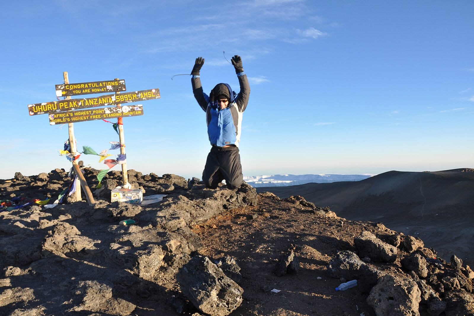 A climber celebrating with arms raised at the summit sign of Kilimanjaro