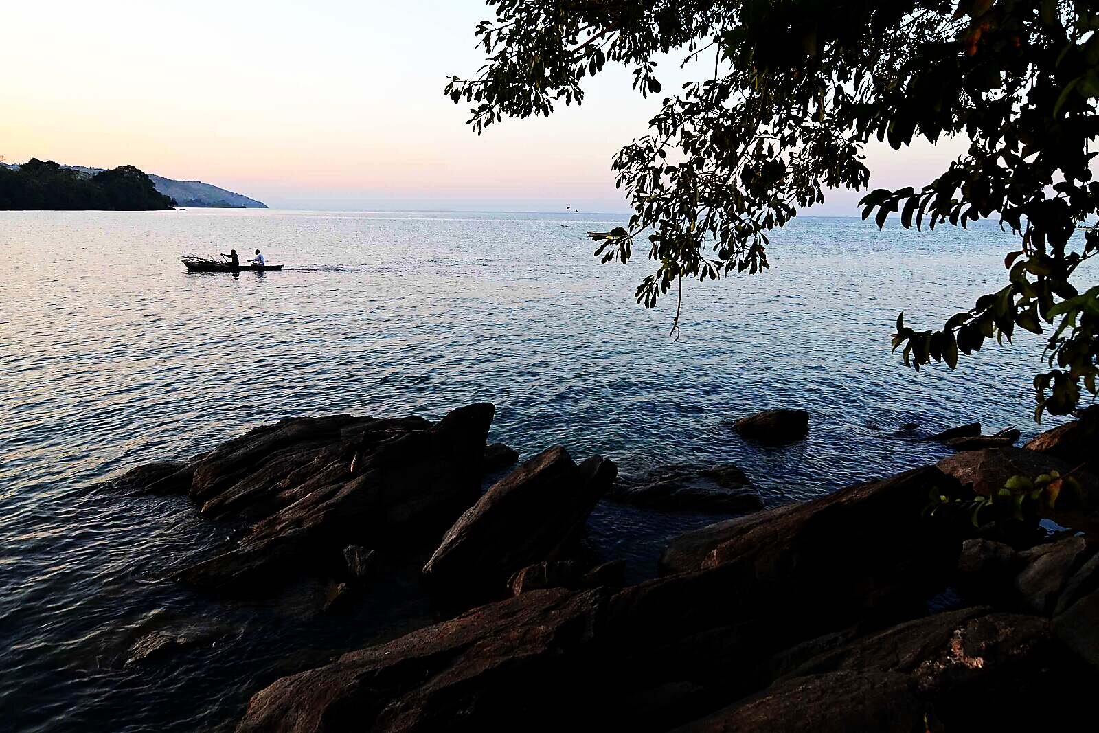 A canoe on mirror-calm Lake Malawi at dusk framed by rocks and trees