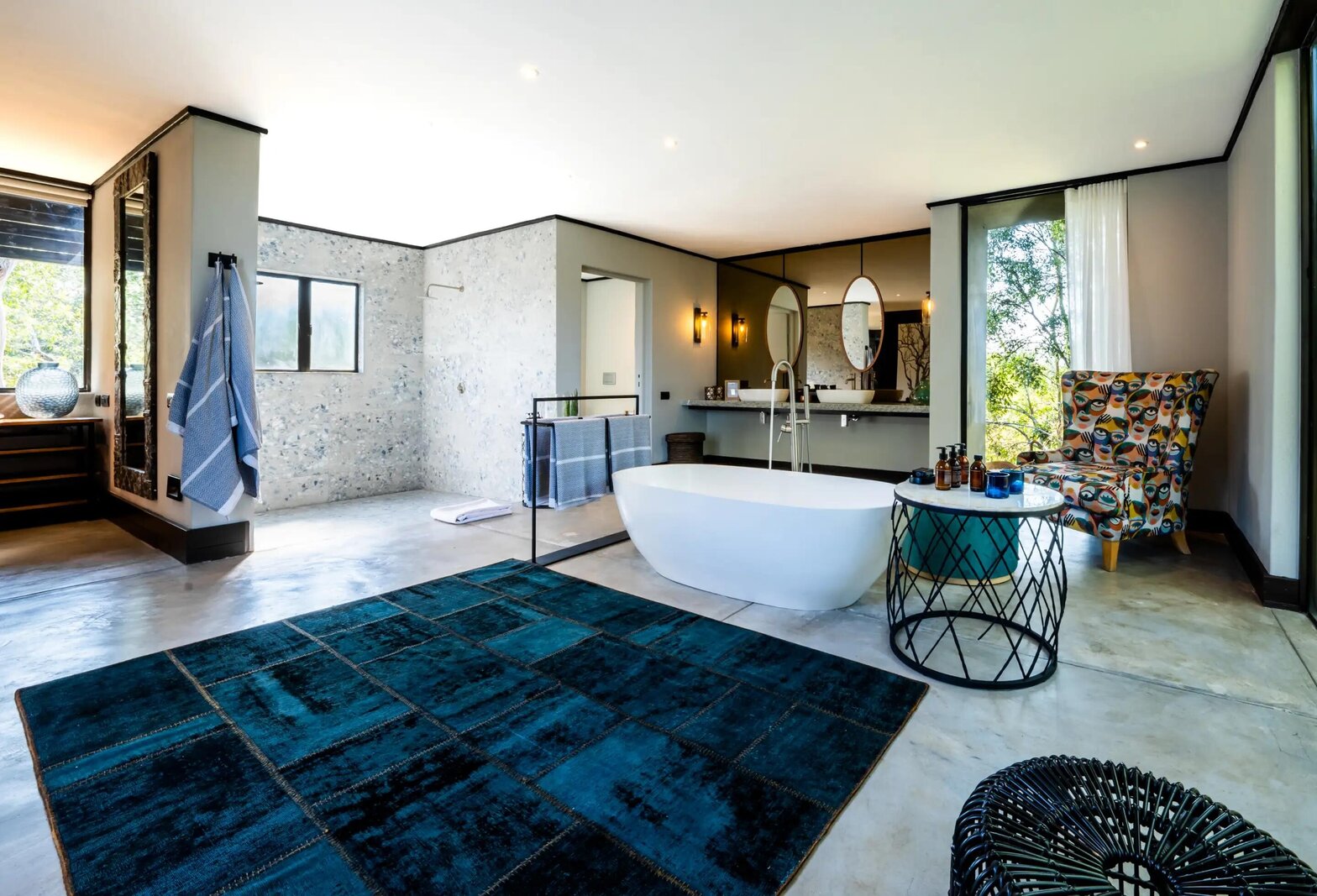 A Silvan Safari suite bathroom with a freestanding tub on a blue patterned rug and floor-to-ceiling glass onto the bush