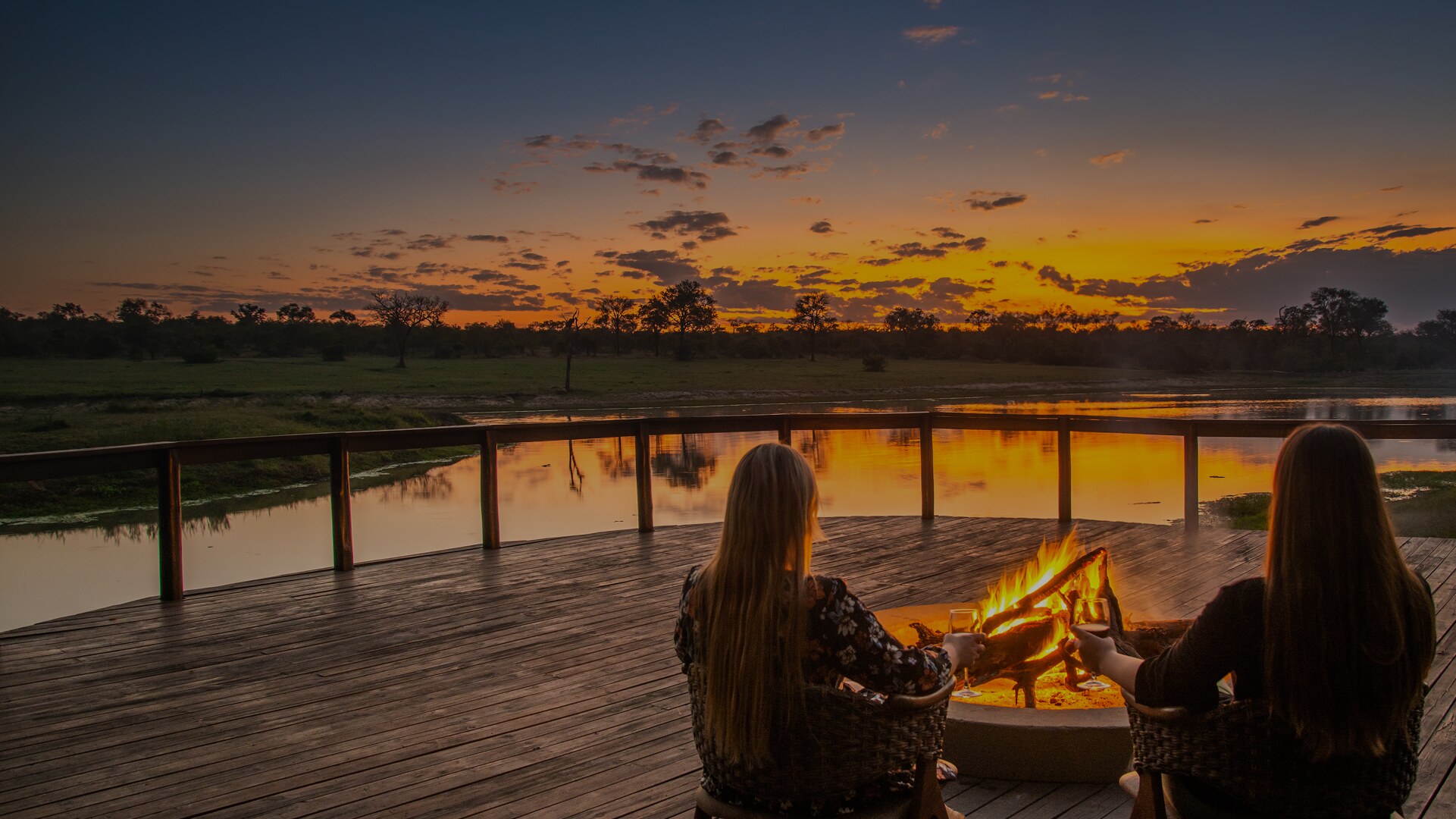 Two guests at a fire pit on the Arathusa Safari Lodge deck at sunset, looking out over the lodge's permanent waterhole in the northern Sabi Sand