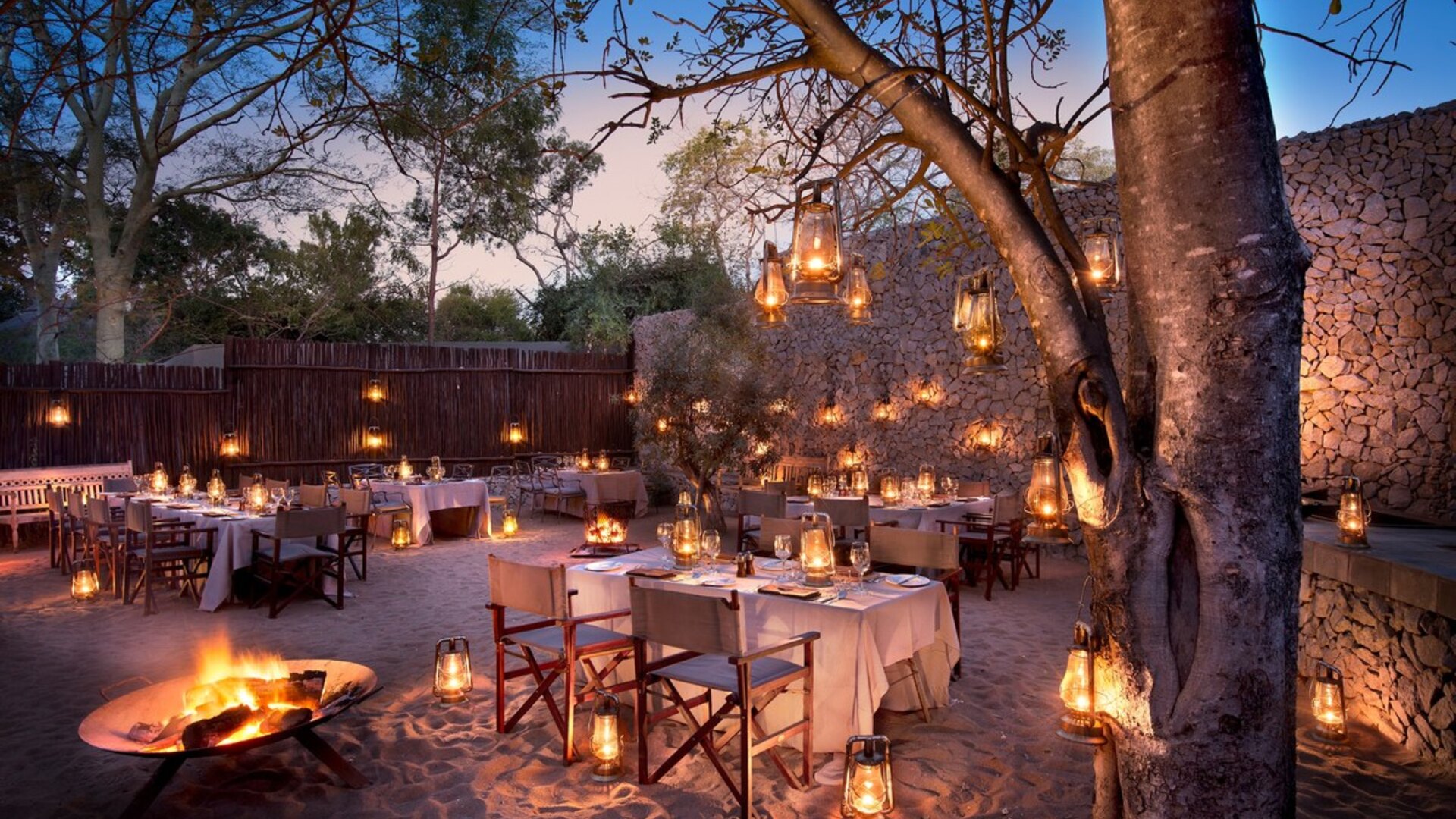 A long candlelit banquet table set under the trees for a corporate group at a luxury African safari lodge at dusk