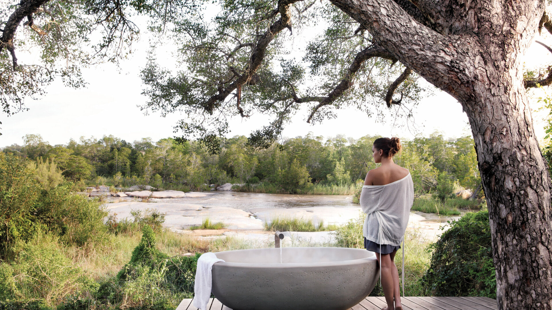 A guest at a Londolozi Private Granite Suite standing beside an outdoor stone bath overlooking the Sand River at golden hour