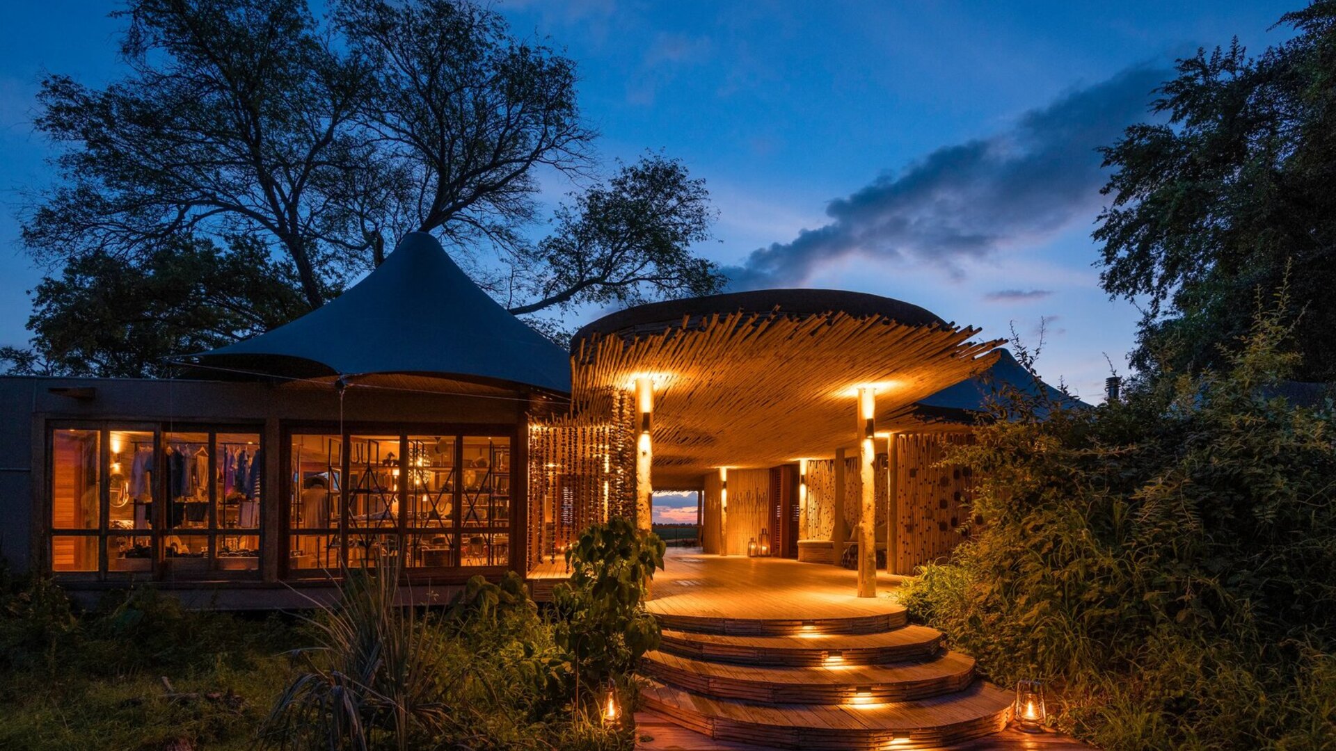 A luxury Okavango Delta camp glowing warmly against a dramatic dusk sky, reflected in the delta waters — an Okavango Delta honeymoon safari with Beyond Africa Safaris