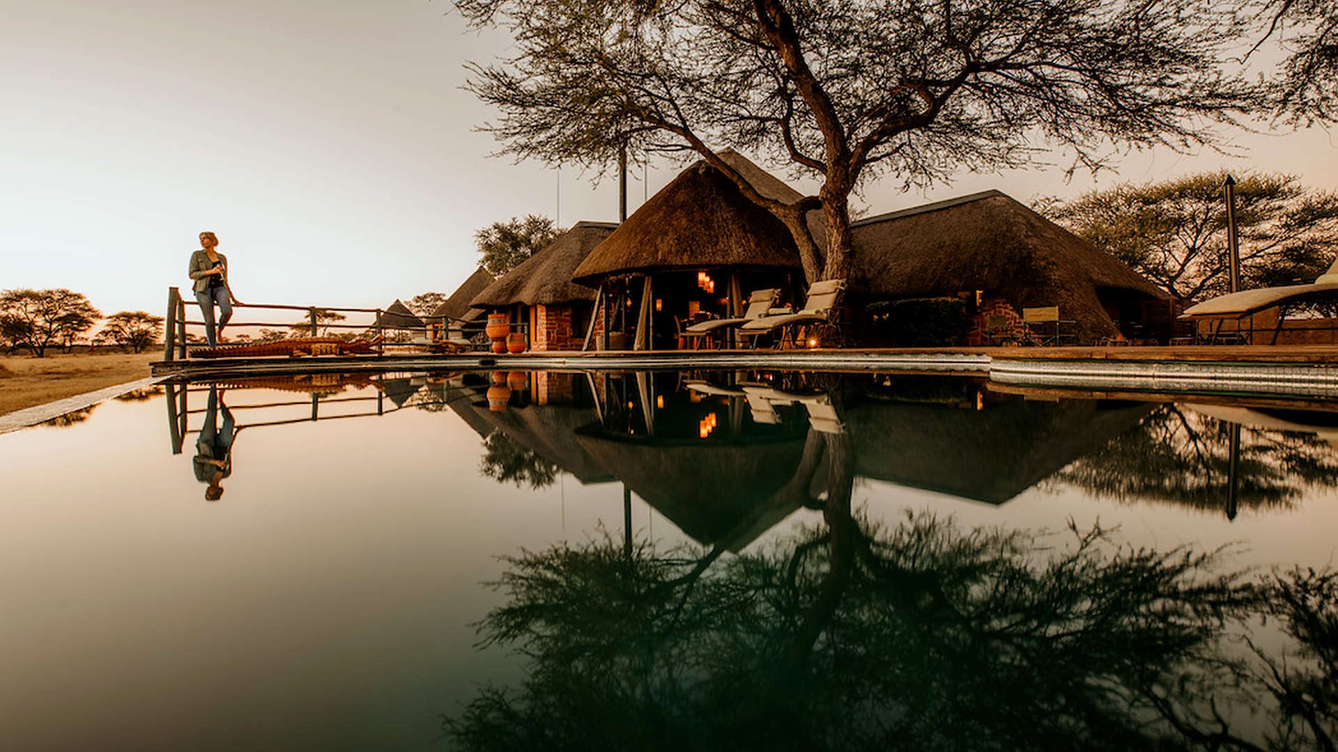 The waterhole and thatched main area of Okonjima Plains Camp reflected at golden hour