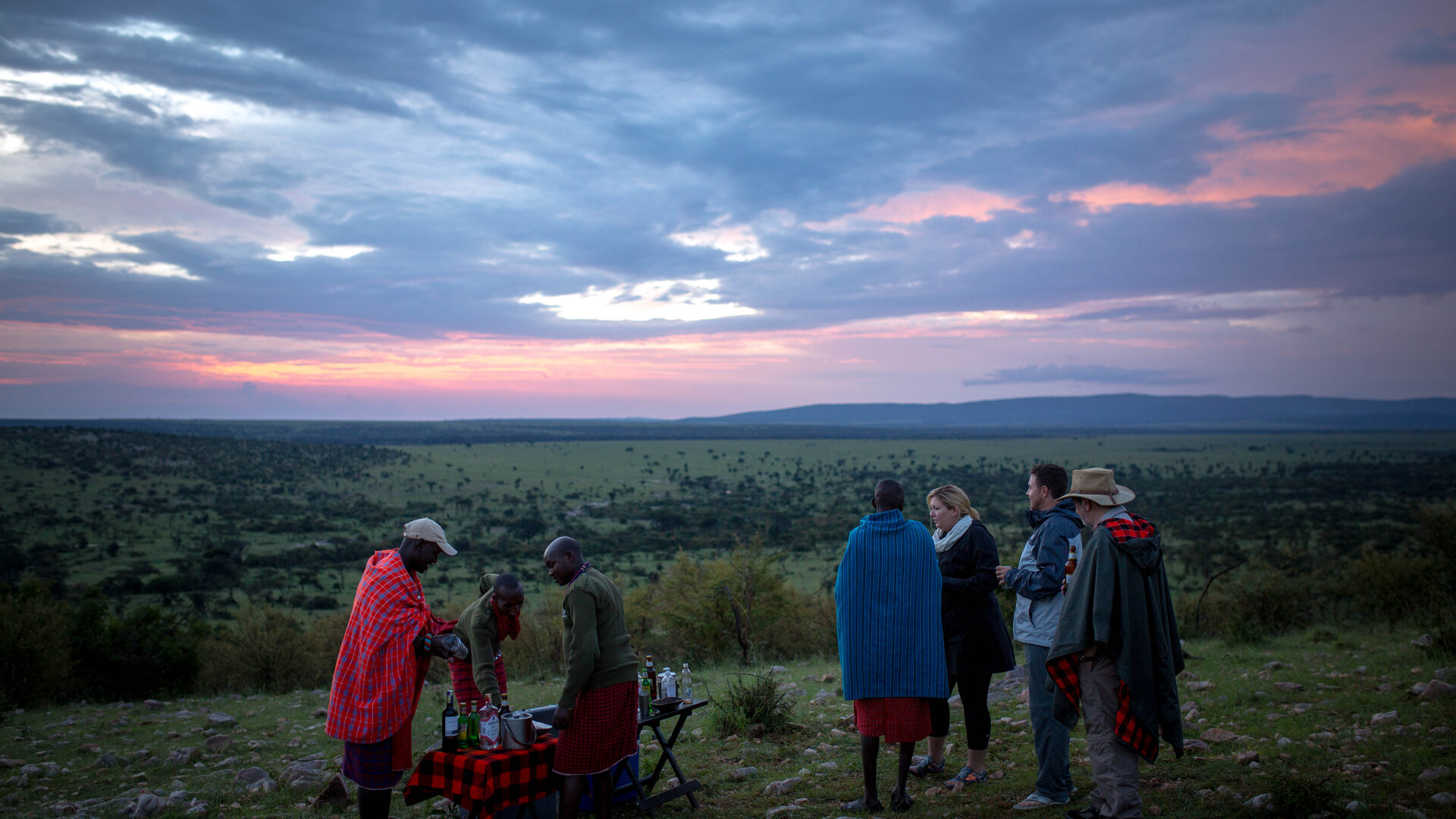 Safari guests enjoying a sundowner with a Maasai guide at dusk on the plains