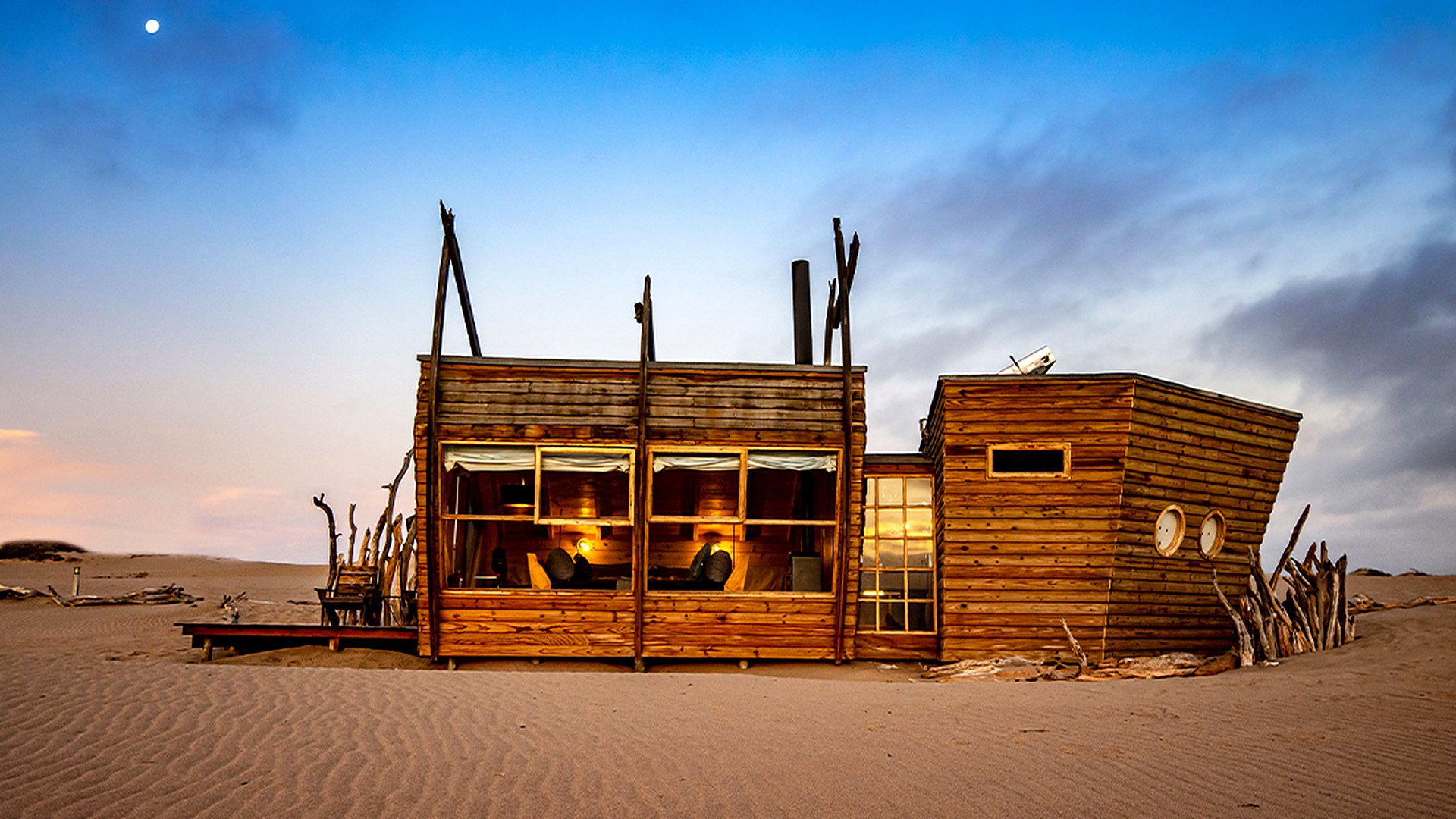A shipwreck-shaped timber cabin rises from the dunes of the Skeleton Coast at golden hour